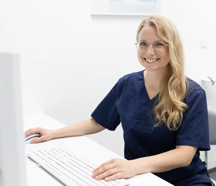Eine Frau in blauer Arbeitskleidung sitzt an einem Schreibtisch vor einem Computer, h&auml;lt eine Hand auf der Maus und l&auml;chelt in die Kamera.