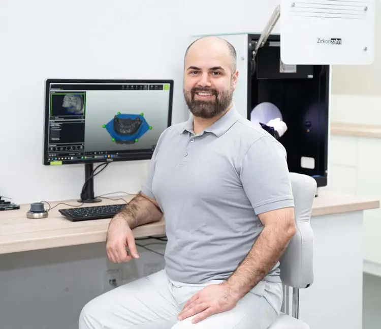 Labortechniker sitzt an einem Scanner mit dem dazugeh&ouml;rigen 3D-Modell am PC und l&auml;chelt in die Kamera